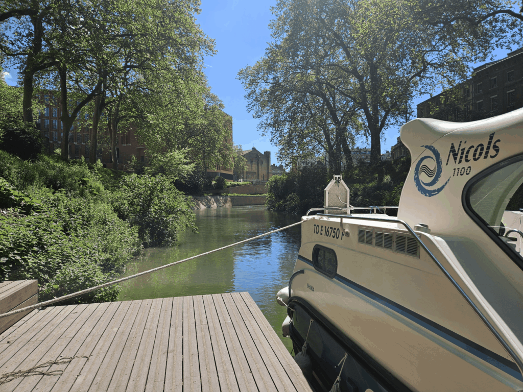Le bateau SHIMA amarré le long d'un ponton de bois, sur un canal bordé de platanes et de bâtiments en brique