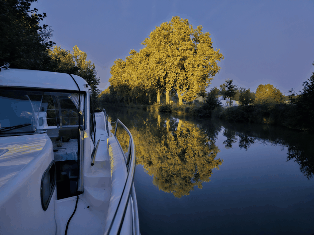 L'avant du bateau SHIMA sur un canal au crépuscule, les arbres dorés se reflétant dans l'eau