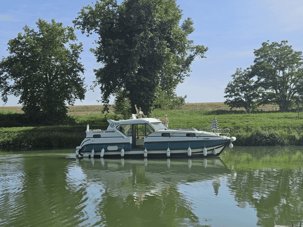 Le bateau SHIMA en navigation sur le canal au milieu des champs, deux personnes sur le pont supérieur