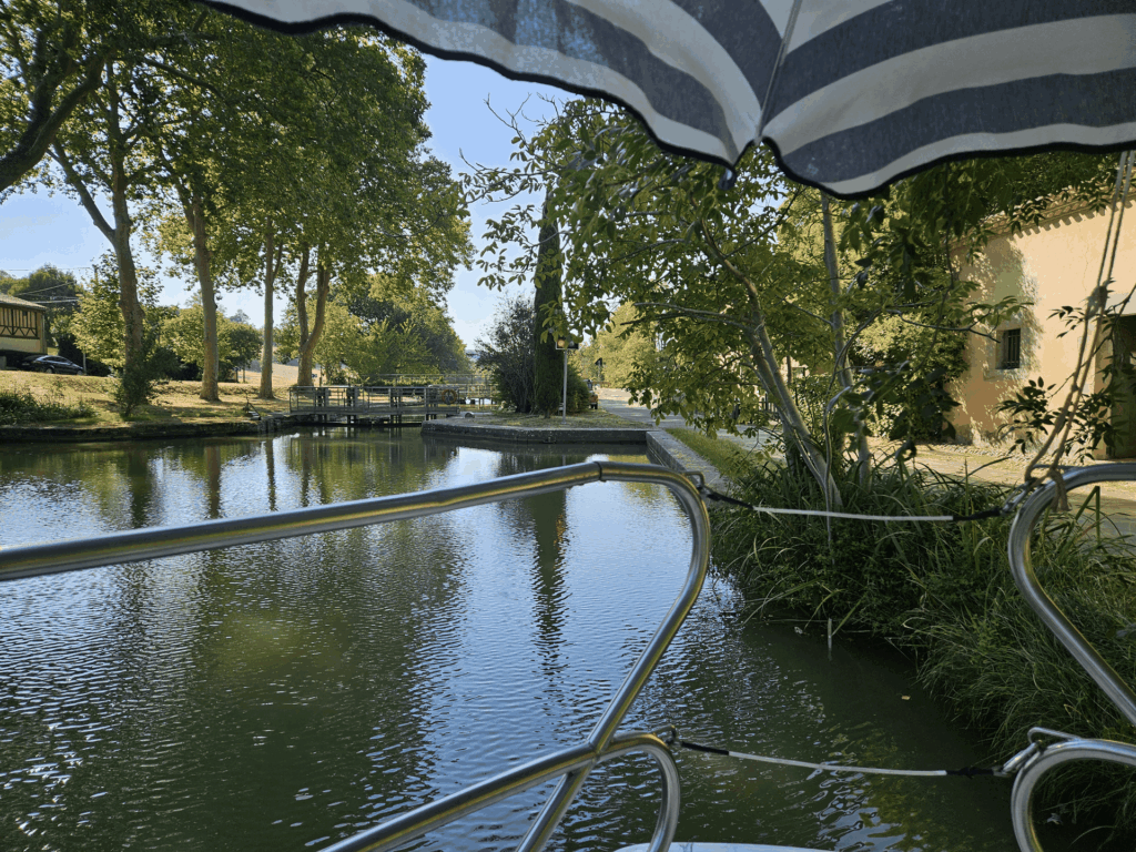 Vue depuis le pont du bateau, sous un parasol rayé, à l'approche d'une écluse bordée de platanes
